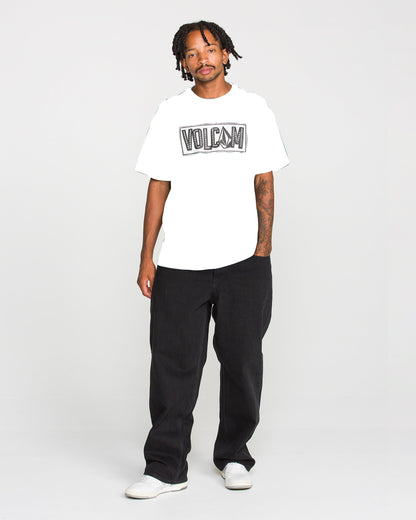 A person stands against a plain background, wearing the Volcom Men's Studded Short Sleeve Tee in white, crafted from heavyweight premium cotton, paired with black pants and white shoes. One arm displays a tattoo, while the tee features a soft-hand screen print that brings subtle style to its classic design.