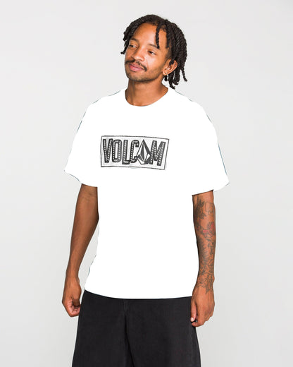 A man with short dreadlocks and forearm tattoos wears the Volcom Men's Studded Short Sleeve Tee, a heavyweight premium cotton T-shirt in white featuring a large soft-hand screen-printed logo. He pairs it effortlessly with loose black shorts while standing against a plain light background, gazing slightly to the side.