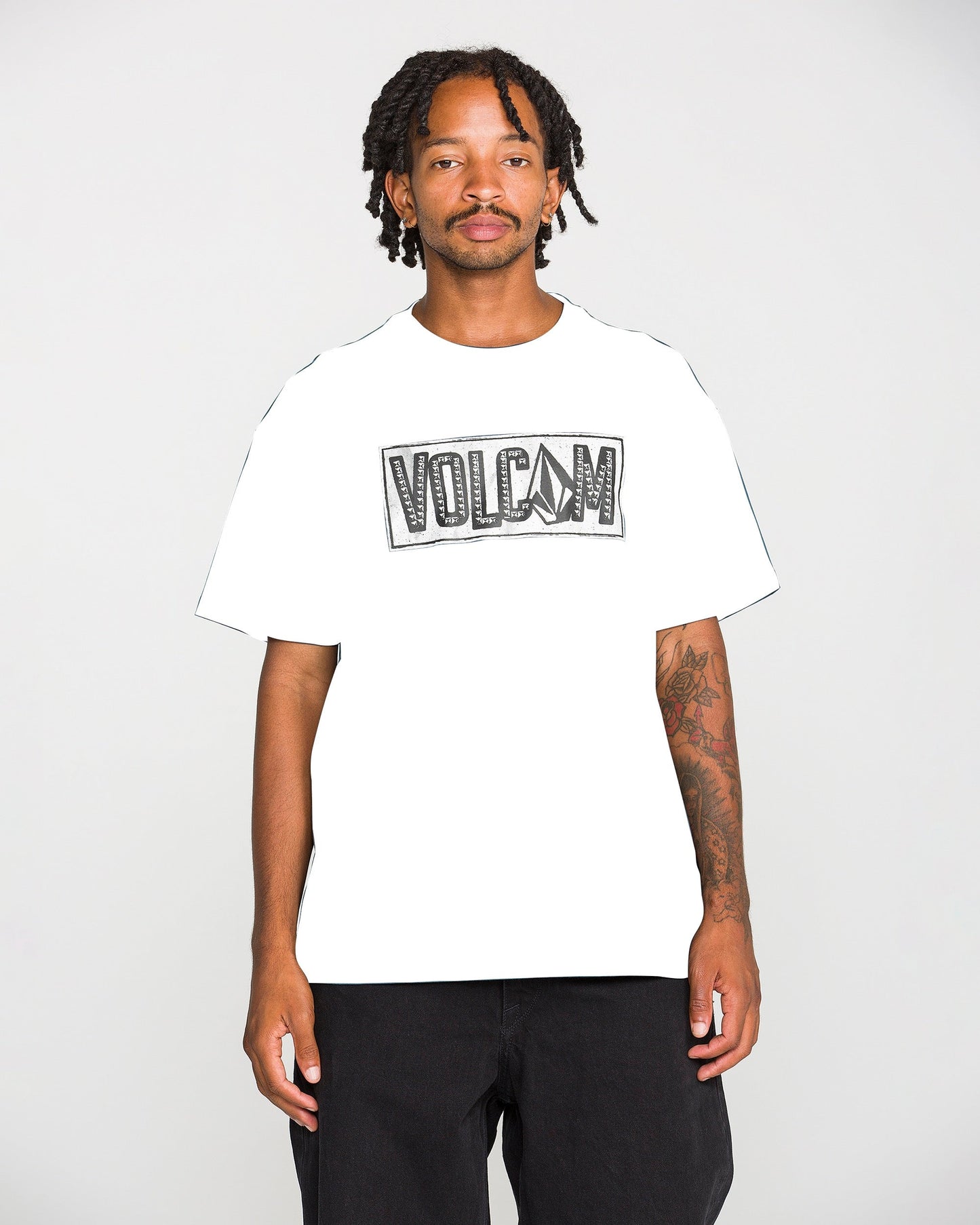 A man with short dreadlocks and a mustache wears the Volcom Men's Studded Short Sleeve Tee, crafted from heavyweight premium cotton and featuring a soft-hand screen print. He pairs it with black pants and stands against a plain light background.