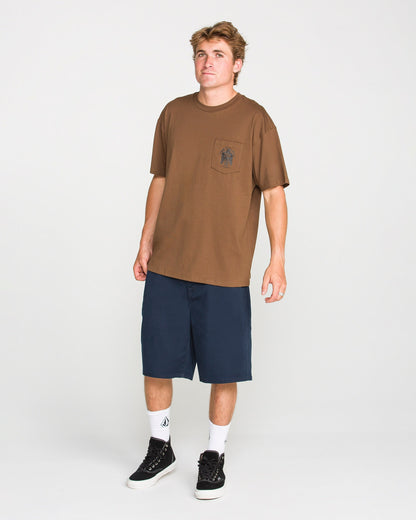 A young man stands against a plain white background wearing Volcom’s Men's Troubador Short Sleeve Tee in brown, crafted from heavyweight ringspun cotton with a practical chest pocket, paired with navy blue shorts, white socks, and black sneakers for a laid-back look.