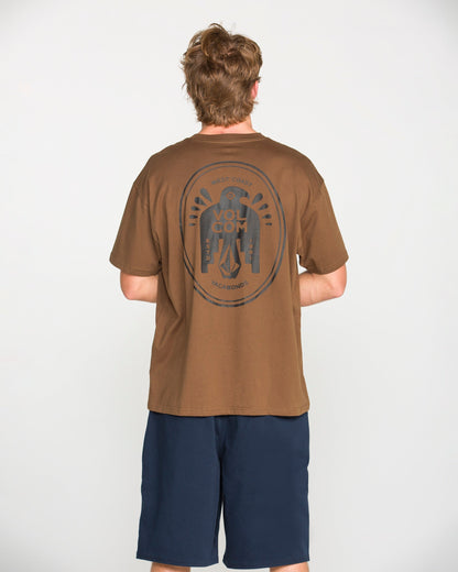 A person with short, light brown hair is standing facing away, wearing the Volcom Men's Troubador Short Sleeve Tee—a brown heavyweight ringspun cotton T-shirt featuring a large circular graphic on the back—paired with navy blue shorts, against a plain white background.