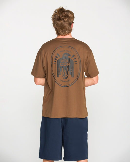 A person with short, light brown hair is standing facing away, wearing the Volcom Men's Troubador Short Sleeve Tee—a brown heavyweight ringspun cotton T-shirt featuring a large circular graphic on the back—paired with navy blue shorts, against a plain white background.