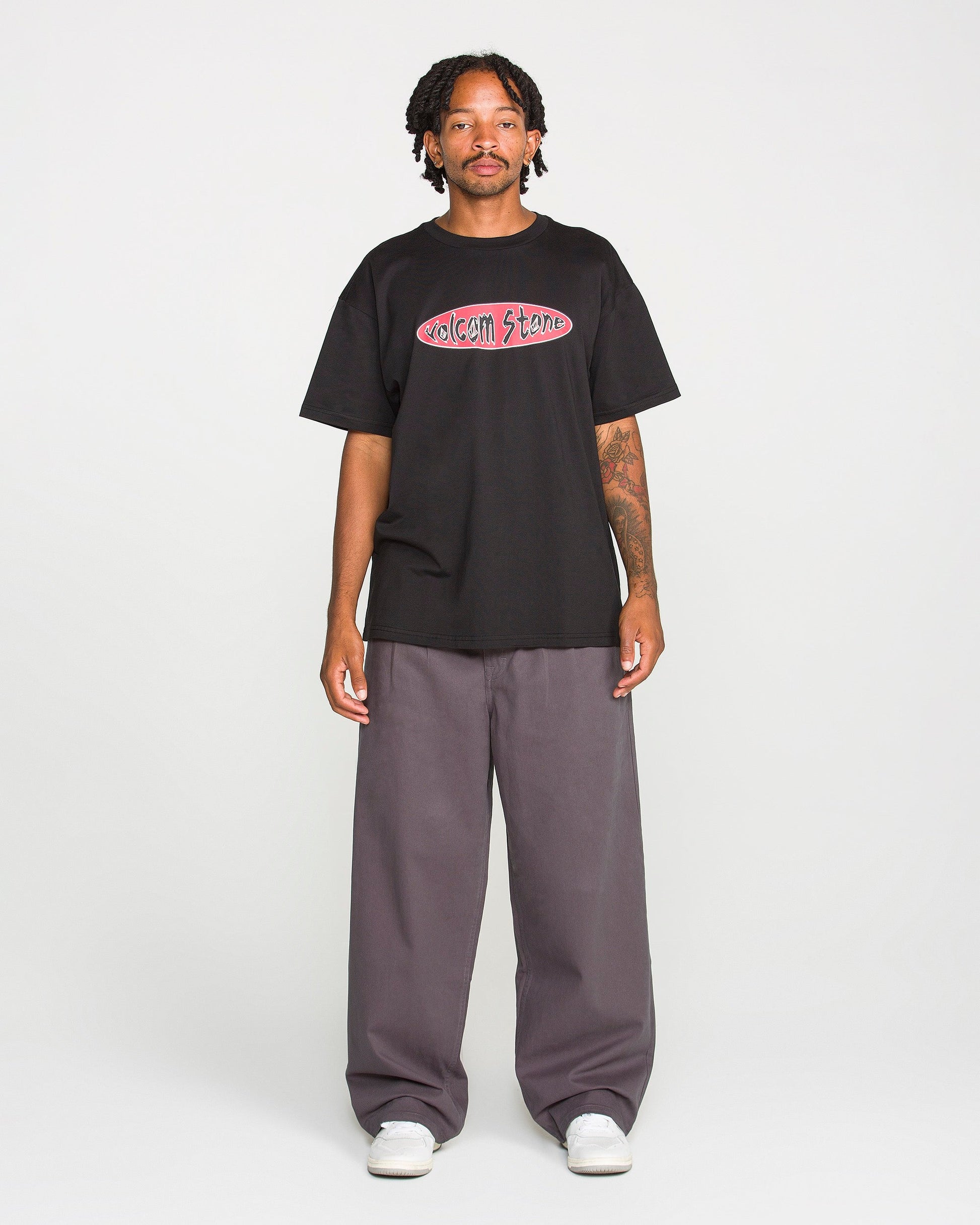 A person stands against a plain background, wearing the Volcom Men's Scratch Stone Short Sleeve Tee in black, made from heavyweight ringspun cotton. They pair it with wide gray pants and white shoes. The individual has short dreadlocks and visible tattoos on one arm.