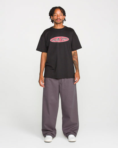 A person stands against a plain background, wearing the Volcom Men's Scratch Stone Short Sleeve Tee in black, made from heavyweight ringspun cotton. They pair it with wide gray pants and white shoes. The individual has short dreadlocks and visible tattoos on one arm.