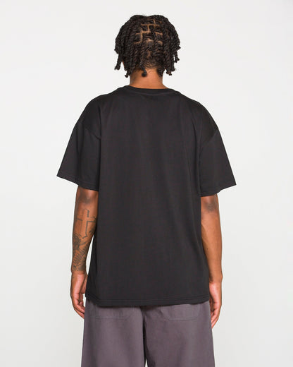 Back view of a person with dark braided hair wearing the Volcom Men's Scratch Stone Short Sleeve Tee in plain black and grey shorts. A large cross tattoo is visible on their right forearm. They are standing against a plain, light grey background.