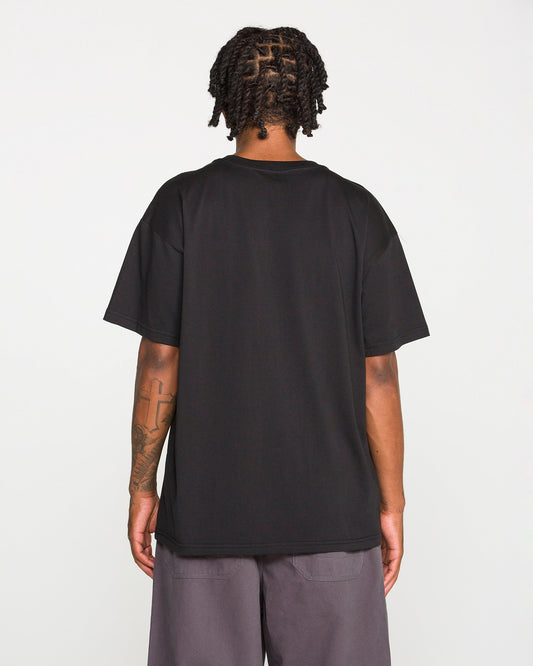 Back view of a person with dark braided hair wearing the Volcom Men's Scratch Stone Short Sleeve Tee in plain black and grey shorts. A large cross tattoo is visible on their right forearm. They are standing against a plain, light grey background.