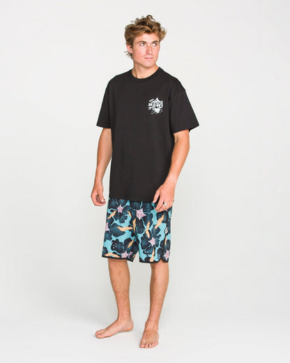 A barefoot person stands on a plain background, wearing the Volcom Men's Concrete Jungle Short Sleeve Tee—a black crew neck made from heavyweight ringspun cotton with a small white chest graphic—paired with blue patterned shorts.