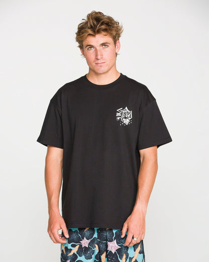 A young man with tousled hair stands against a plain white background, looking at the camera with a neutral expression. He is wearing the Volcom Men's Concrete Jungle Short Sleeve Tee, crafted from heavyweight ringspun cotton, paired with patterned shorts. The crew neck t-shirt features a bold graphic that adds an urban edge to his relaxed look.