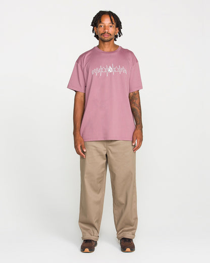 A man with short dreadlocks wears the Volcom Men's Repeater Short Sleeve Tee in a mauve hue, featuring a loose fit and crafted from heavyweight ringspun cotton. He pairs it with beige pants and brown shoes, standing facing forward against a plain white background with a neutral expression.