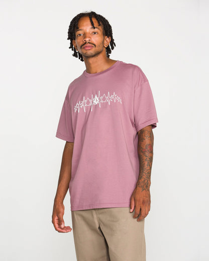 A man with short dreadlocks wears the Volcom Men's Repeater Short Sleeve Tee—a mauve loose-fit t-shirt with a white abstract design—paired with beige pants, standing against a plain white background.