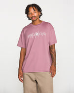 A man wearing the Volcom Men's Repeater Short Sleeve Tee in mauve, featuring a white abstract graphic across the chest and paired with beige pants, stands against a plain white background. This loose fit crew neck tee is crafted from heavyweight ringspun cotton for exceptional comfort and durability.