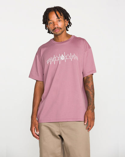 A man wearing the Volcom Men's Repeater Short Sleeve Tee in mauve, featuring a white abstract graphic across the chest and paired with beige pants, stands against a plain white background. This loose fit crew neck tee is crafted from heavyweight ringspun cotton for exceptional comfort and durability.