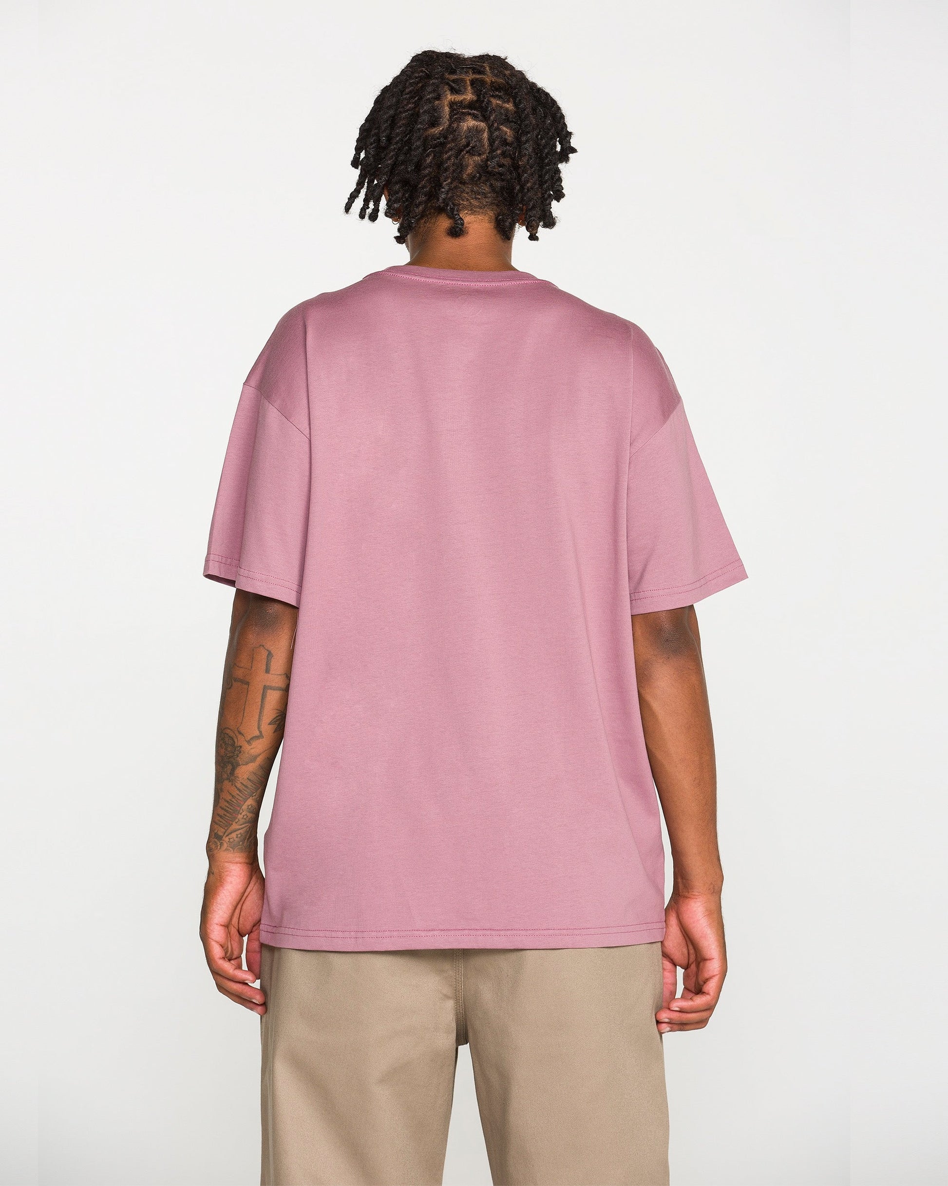 A person with short twisted hair stands facing away from the camera, wearing Volcom's Men's Repeater Short Sleeve Tee in mauve—a loose fit crafted from heavyweight ringspun cotton—paired with beige pants, showcasing the back of the shirt.