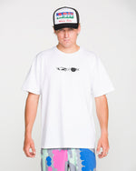 A person wearing a white Volcom Men's Featured Artist Head Check Short Sleeve Tee, crafted from premium ringspun cotton for superior comfort and style and designed by Antonio Carrau, is paired with colorful abstract shorts and a black trucker hat featuring a vibrant graphic design, standing against a plain white background.
