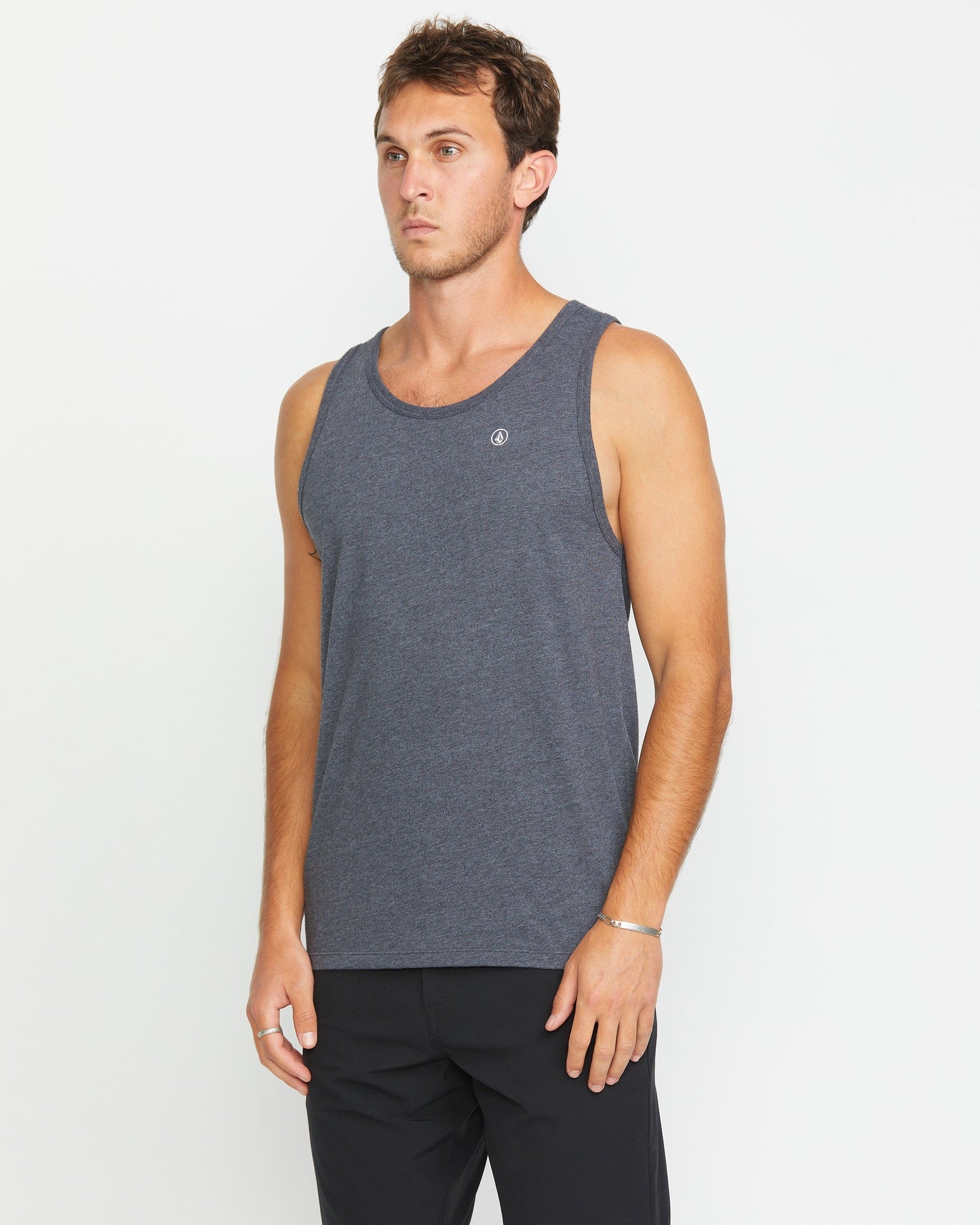 A man wears the Volcom Men's Solid Heather Tank in a modern fit with dark black heather pants, standing against a white background and looking to the side with a neutral expression.