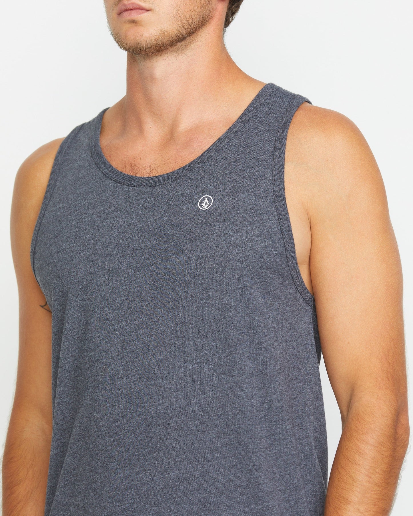 A man stands against a white background wearing the Volcom Men's Solid Heather Tank in dark black heather, featuring a small white circular logo on the chest. His face is partially out of frame.