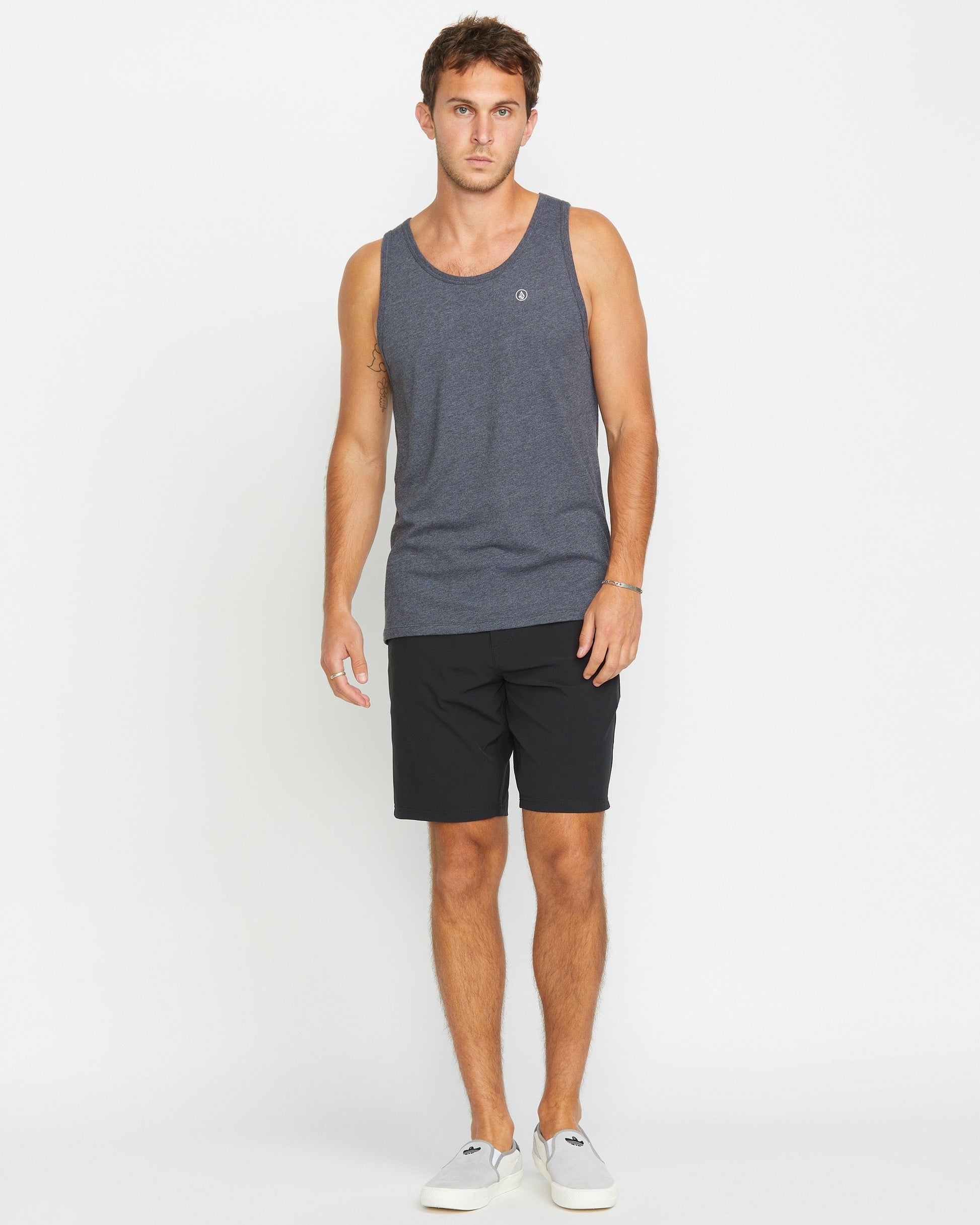 A man with short brown hair and a neutral expression wears the Volcom Men's Solid Heather Tank, paired with black shorts and light gray slip-on shoes, standing against a plain white background.