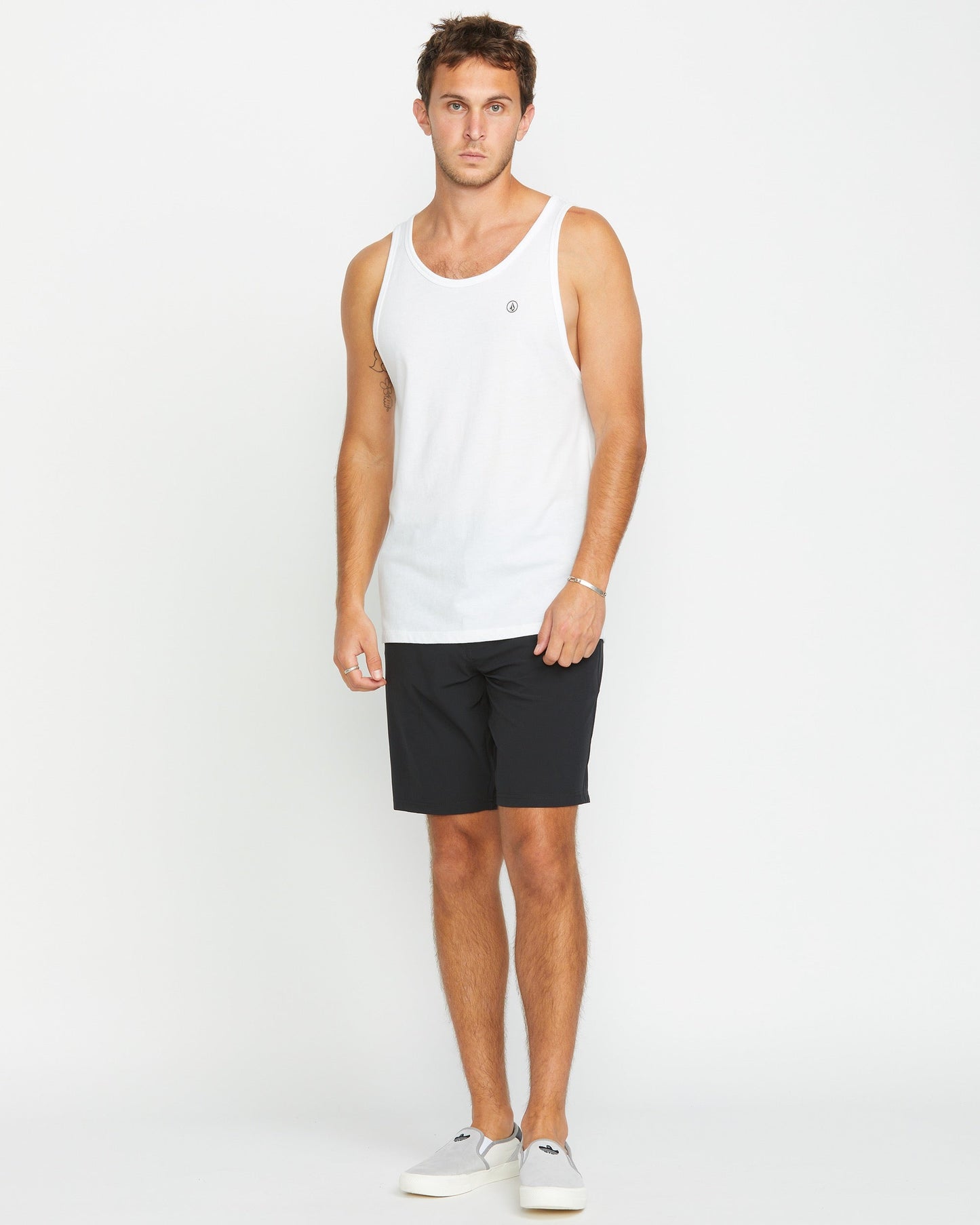 A man faces forward against a plain white background, wearing the Volcom Men's Solid Heather Tank in white with black shorts and white slip-on shoes, highlighting a clean, modern fit.