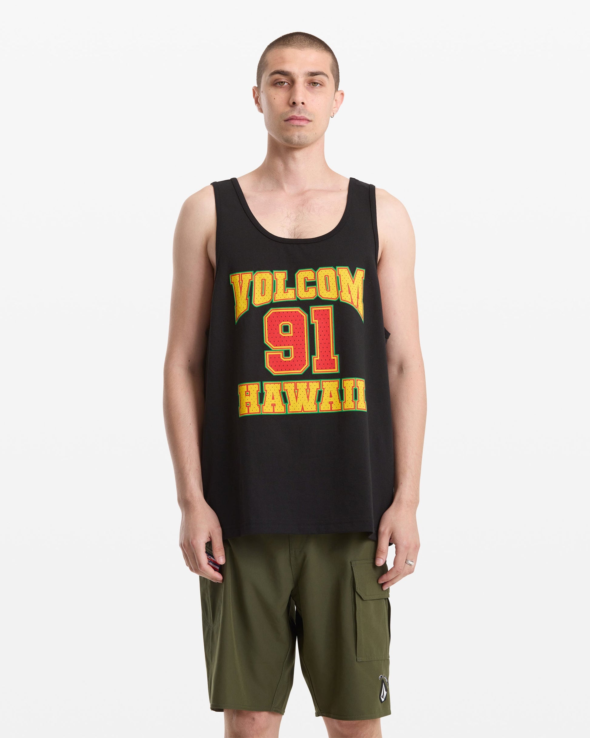 A man with short hair wears the Volcom Men's Swish Tank, a loose black top with "Volcom 91 Hawaii" in yellow and red, paired with olive cargo shorts, standing against a plain white background.