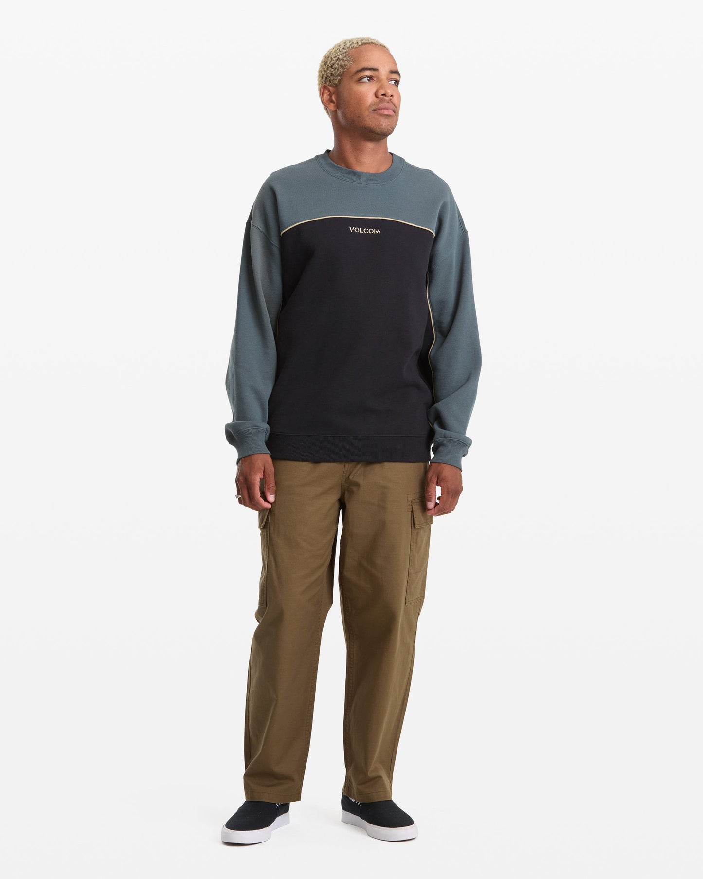 A person with short blond hair wears the Volcom Men's 2K Stone Crew sweatshirt, khaki pants, and black slip-on shoes, standing against a plain white background and looking slightly to the side.