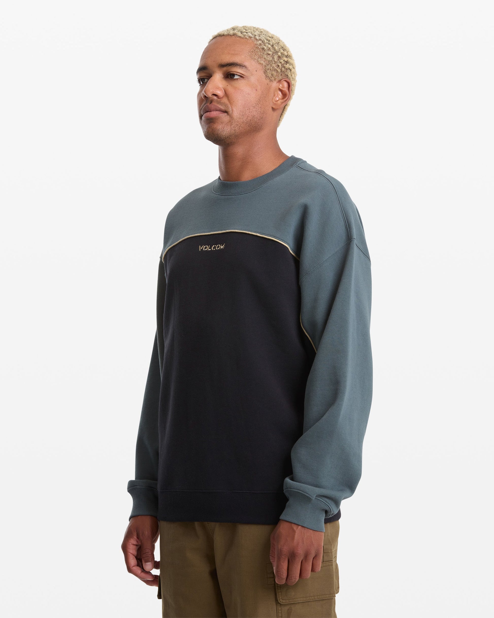 A person with short, bleached hair wears the Volcom Men's 2K Stone Crew sweatshirt in blue and black with tan pants, standing against a plain white background.