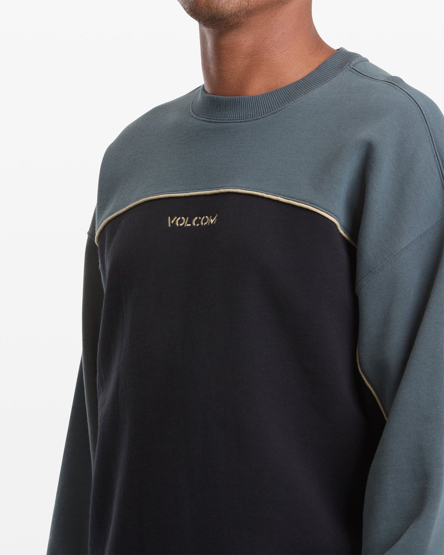 A person wears the Volcom Men's 2K Stone Crew, a color-blocked sweatshirt made from heavyweight fleece with dark blue sleeves and lower half, black chest, gold VOLCOM embroidery, and a bold gold accent line.