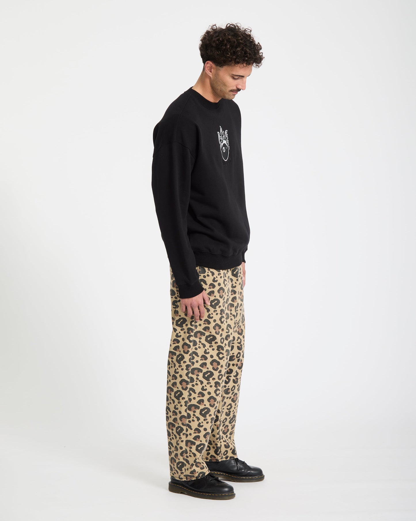 A man stands against a plain white background, wearing the Volcom Men's Featured Artist Seth Conboy Crew sweatshirt, loose tan leopard print pants, and black shoes. He looks down with his arms at his sides.