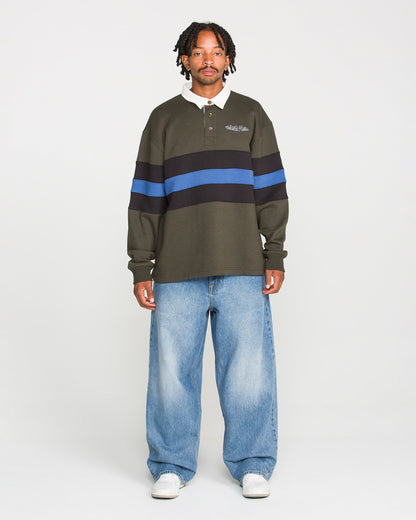 A man stands facing forward against a plain white background, wearing the Volcom Men's Scratch Stone Fleece Polo—featuring a boxy fit in dark green with blue and black stripes—paired with baggy light blue jeans and white sneakers.
