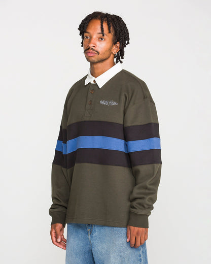 A person wearing the Volcom Men's Scratch Stone Fleece Polo—a dark green long-sleeve shirt with black and blue horizontal stripes in a boxy fit—and light blue jeans stands against a plain white background.