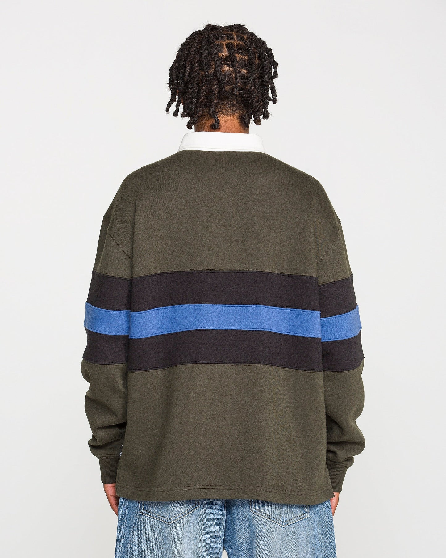 A person with short twists stands facing away, wearing the Volcom Men's Scratch Stone Fleece Polo in a boxy fit olive green with black and blue horizontal stripes, crafted from recycled polyester fleece, paired with light blue jeans against a plain white background.