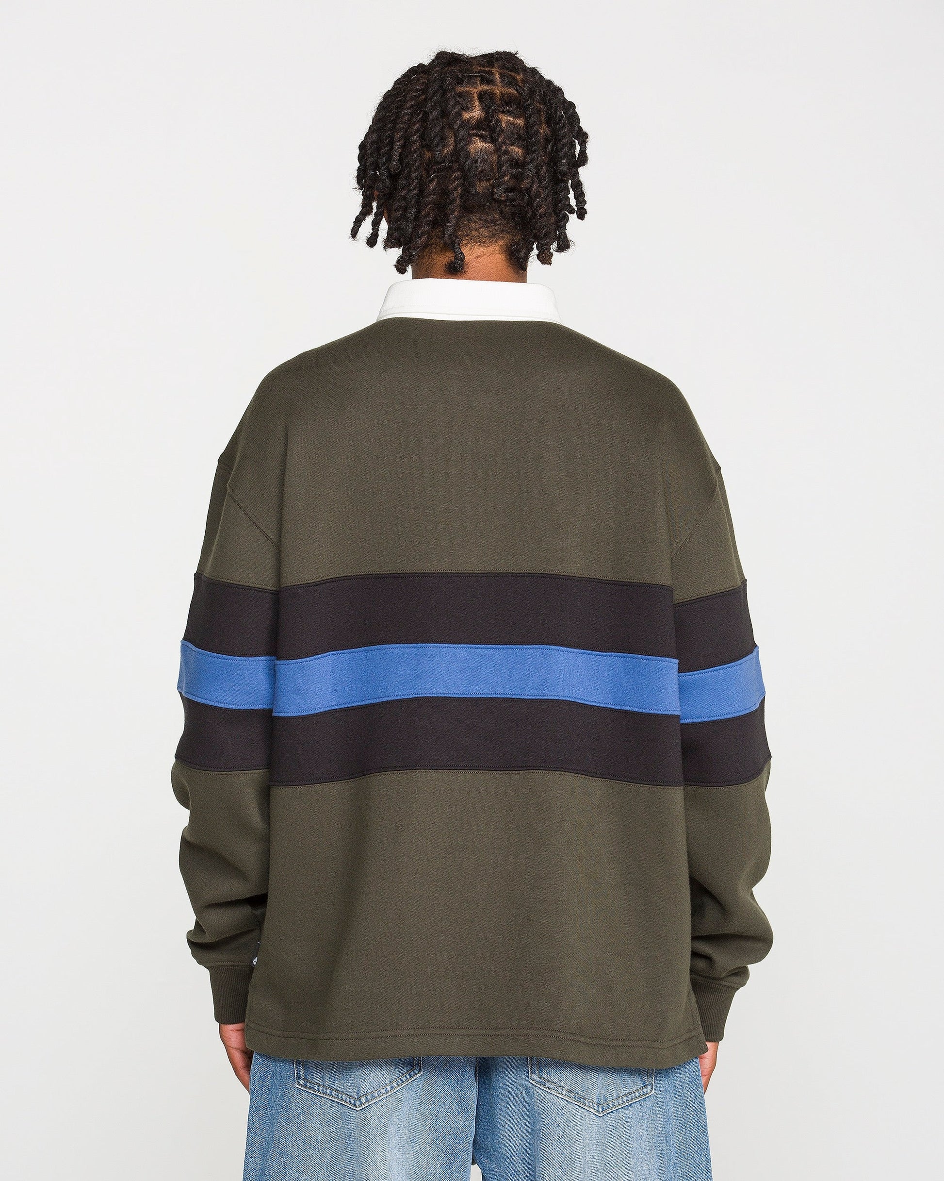 A person with short twists stands facing away, wearing the Volcom Men's Scratch Stone Fleece Polo in a boxy fit olive green with black and blue horizontal stripes, crafted from recycled polyester fleece, paired with light blue jeans against a plain white background.