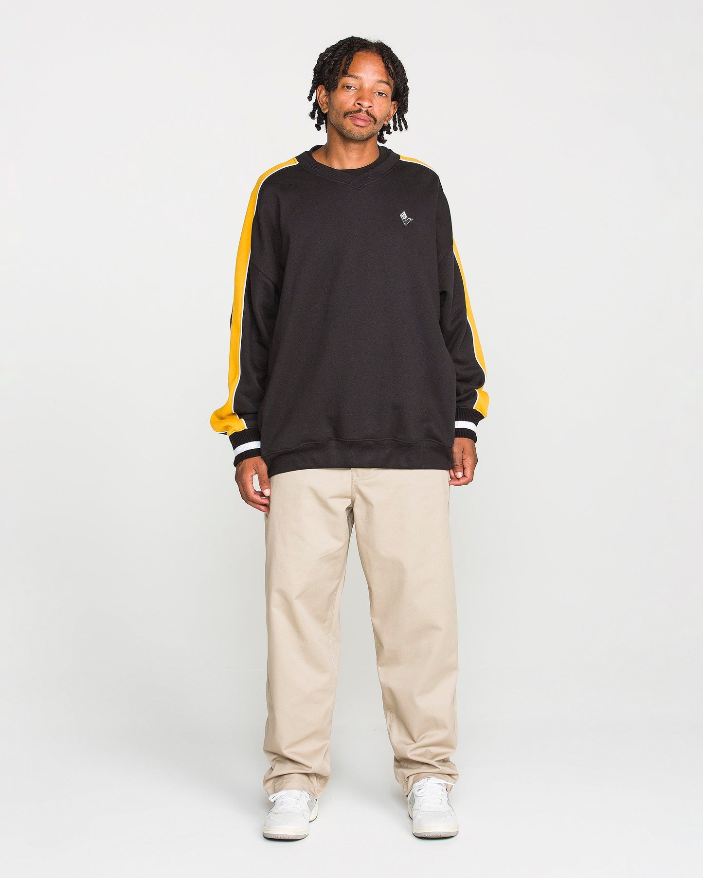 A person stands facing forward, wearing the Volcom Men's Flying Hi Crew—a black sweatshirt with yellow colorblocked sleeves made from brushed-back fleece featuring REPREVE recycled polyester—paired with beige pants and white sneakers against a plain white background.
