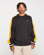 A man with short dreadlocks wears the Volcom Men's Flying Hi Crew, a black sweatshirt with yellow and white colorblocked sleeves crafted from brushed-back fleece with REPREVE recycled polyester, paired with beige pants and standing against a plain light background.