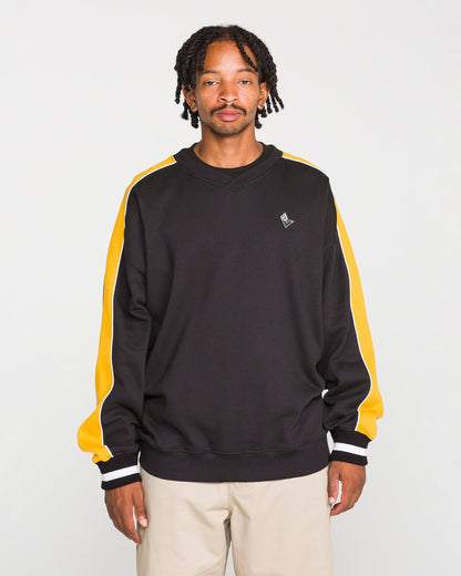 A man with short dreadlocks wears the Volcom Men's Flying Hi Crew, a black sweatshirt with yellow and white colorblocked sleeves crafted from brushed-back fleece with REPREVE recycled polyester, paired with beige pants and standing against a plain light background.