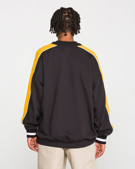 A person is facing away from the camera, wearing the Volcom Men's Flying Hi Crew—a black sweatshirt with colorblocked sleeves featuring yellow and white stripes—paired with light-colored pants, set against a plain white background.