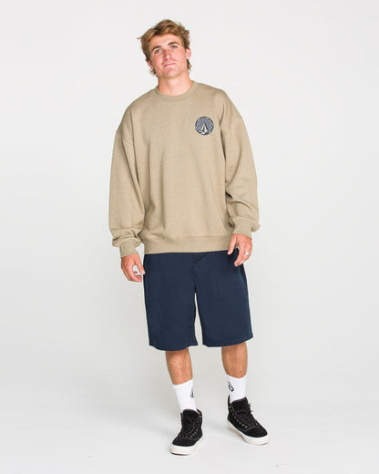 A young man stands against a plain white background, wearing the Volcom Men's Dirty Work Crew—an easygoing beige (khaki) brushed-back fleece sweatshirt with a small circular logo—paired with navy blue shorts, white socks, and black sneakers. The loose fit of the Volcom sweatshirt enhances his relaxed expression as he looks at the camera.