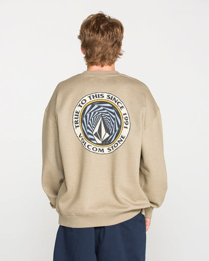A person faces away, wearing the Volcom Men's Dirty Work Crew in a khaki tone. The sweatshirt features a loose fit and comfortable brushed-back fleece. On the back, a circular logo reading "TRUE TO THIS SINCE 1991 VOLCOM STONE" surrounds an abstract geometric design, adding distinctive style to its casual look.