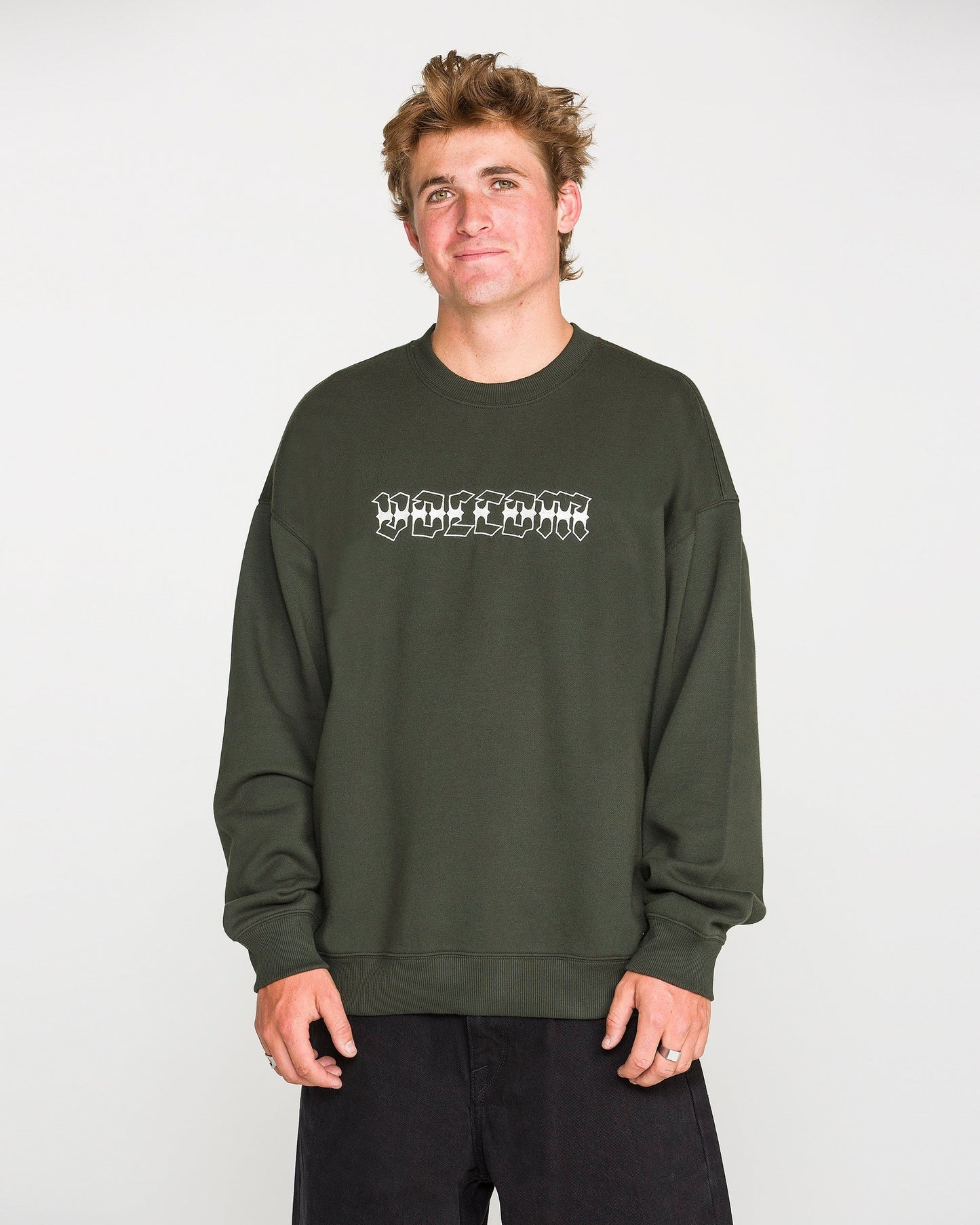 A person with short, tousled hair wears the Volcom Men's Dirty Work Crew— a green, loose fit fleece crew sweatshirt crafted from recycled polyester—paired with black pants and standing against a plain light background. The sweatshirt features white text for added style and offers both comfort and sustainability.
