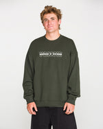 A person with short, tousled hair wears the Volcom Men's Dirty Work Crew— a green, loose fit fleece crew sweatshirt crafted from recycled polyester—paired with black pants and standing against a plain light background. The sweatshirt features white text for added style and offers both comfort and sustainability.