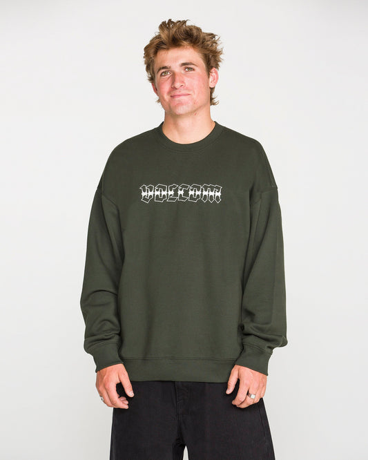 A person with short, tousled hair wears the Volcom Men's Dirty Work Crew— a green, loose fit fleece crew sweatshirt crafted from recycled polyester—paired with black pants and standing against a plain light background. The sweatshirt features white text for added style and offers both comfort and sustainability.