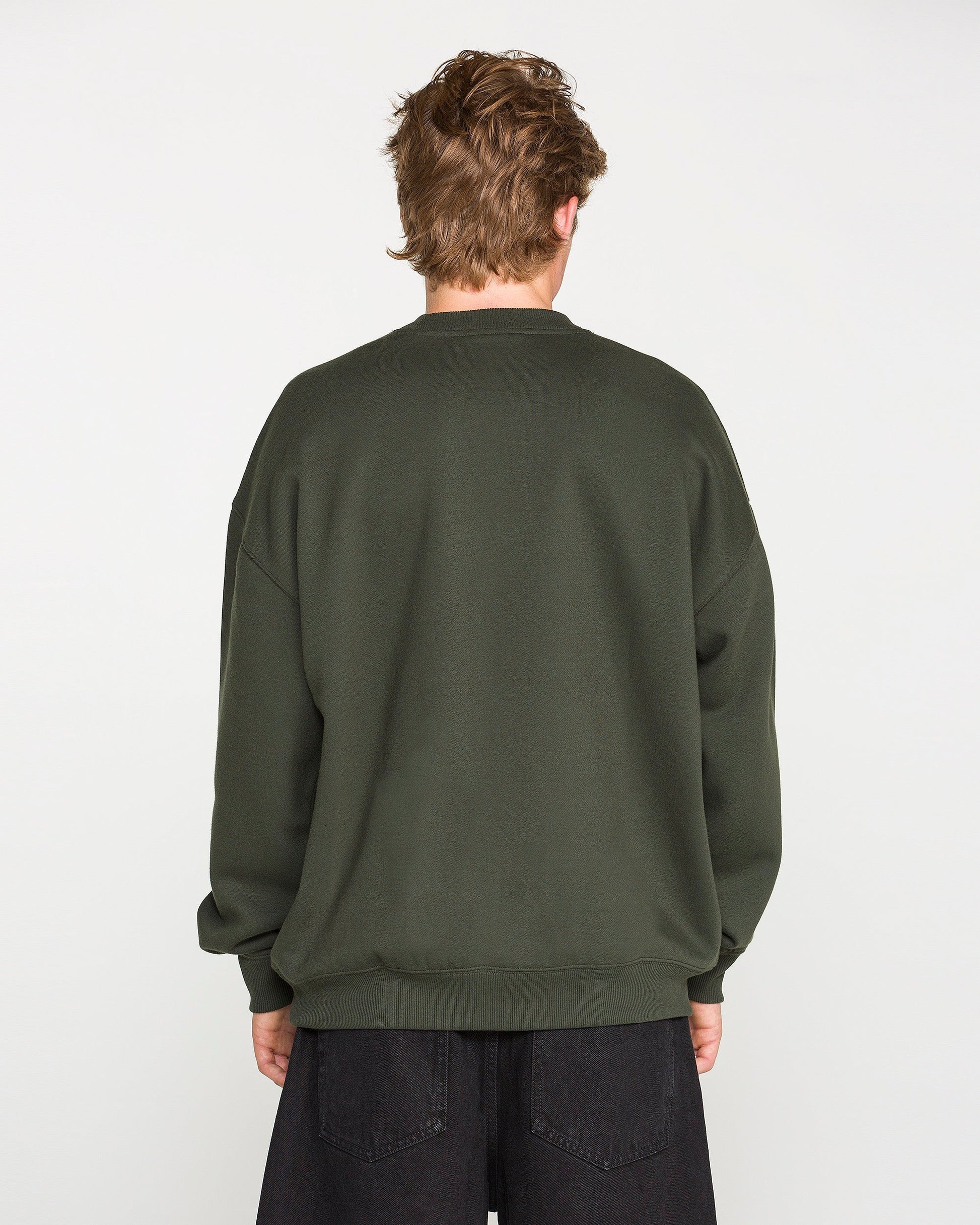 A person with short, light brown hair is facing away from the camera, wearing the Volcom Men's Dirty Work Crew—an oversized green sweatshirt crafted from recycled polyester—paired with black pants against a plain white background.