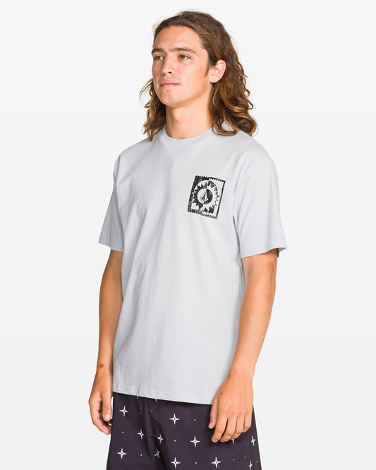 A person with long brown hair wears the Volcom Men's Burner Short Sleeve Tee with a black graphic soft-hand print on the left chest and dark shorts with a white cross pattern, standing against a plain white background.