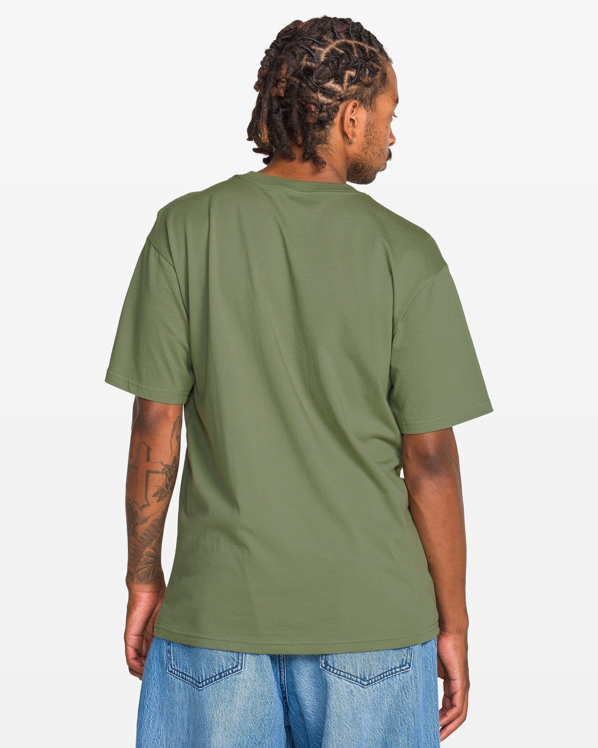 A man with short dreadlocks, dressed in Volcom’s Men's Be Kind Rewind Short Sleeve Tee and light blue jeans, stands with his back to the camera against a plain white background.