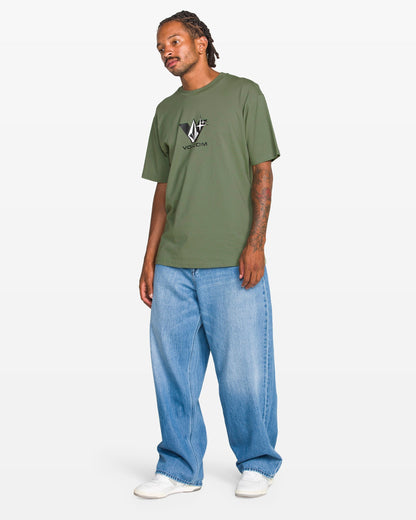 A man in a Volcom Men's Be Kind Rewind Short Sleeve Tee (classic fit, olive green), baggy light blue jeans, and white sneakers stands against a plain white background.