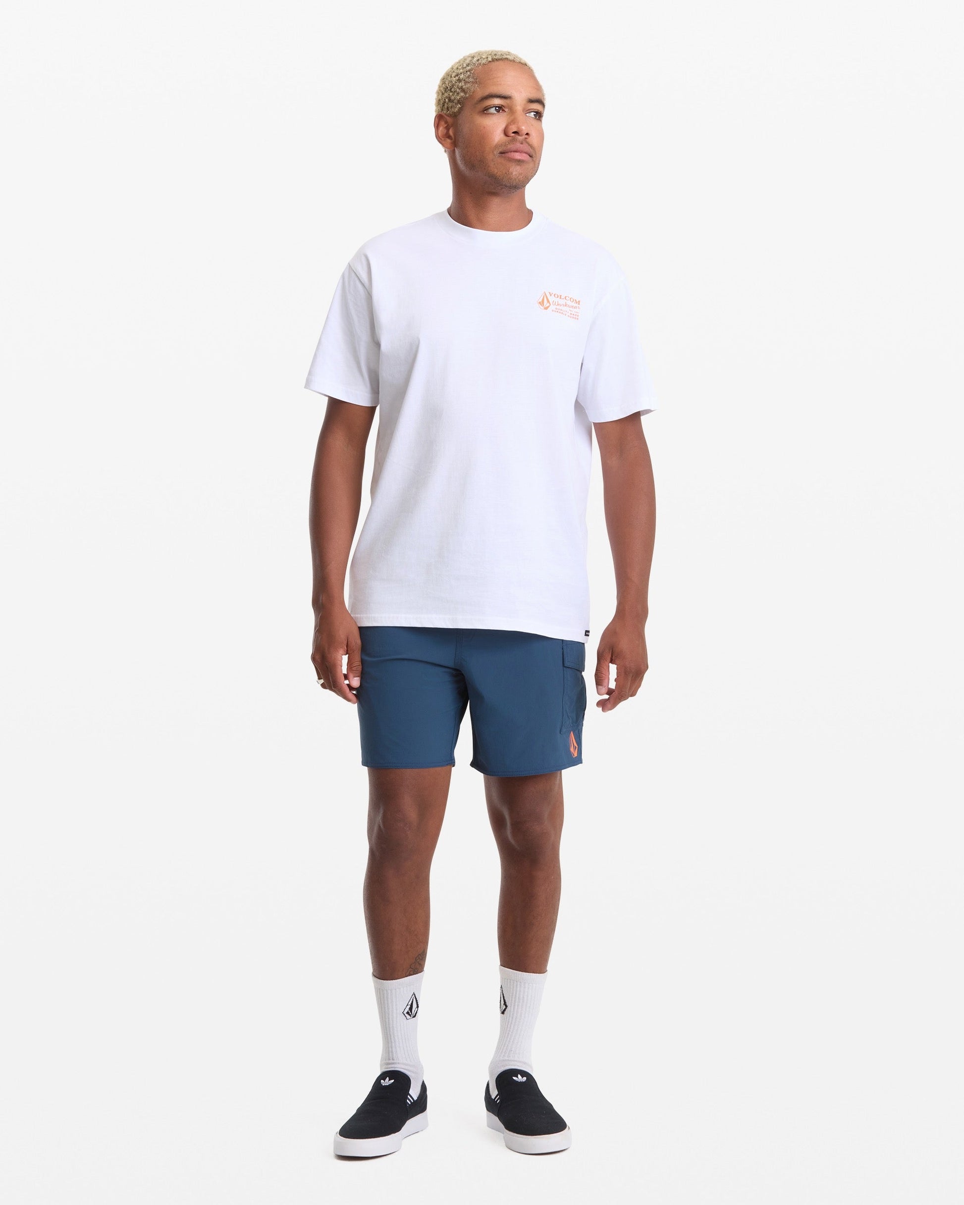 A person stands facing forward against a plain white background wearing a Volcom Men's Workwear Short Sleeve Tee in white, paired with blue shorts, white socks, and black slip-on shoes.