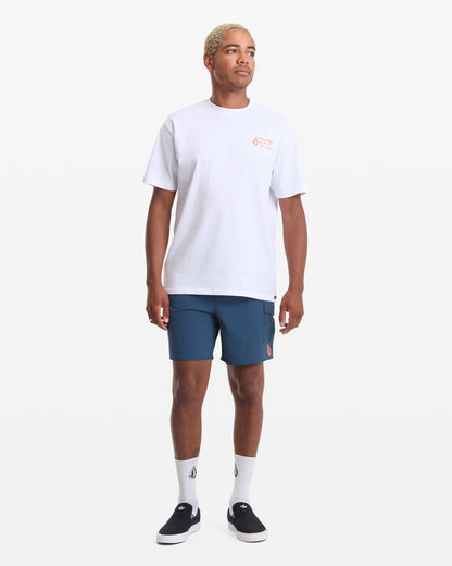 A person stands facing forward against a plain white background wearing a Volcom Men's Workwear Short Sleeve Tee in white, paired with blue shorts, white socks, and black slip-on shoes.