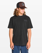 A man wearing the Volcom Men's Proto Type Short Sleeve Tee, blue shorts, and a black Volcom cap stands against a white background, looking down and to the side, channeling effortless style.