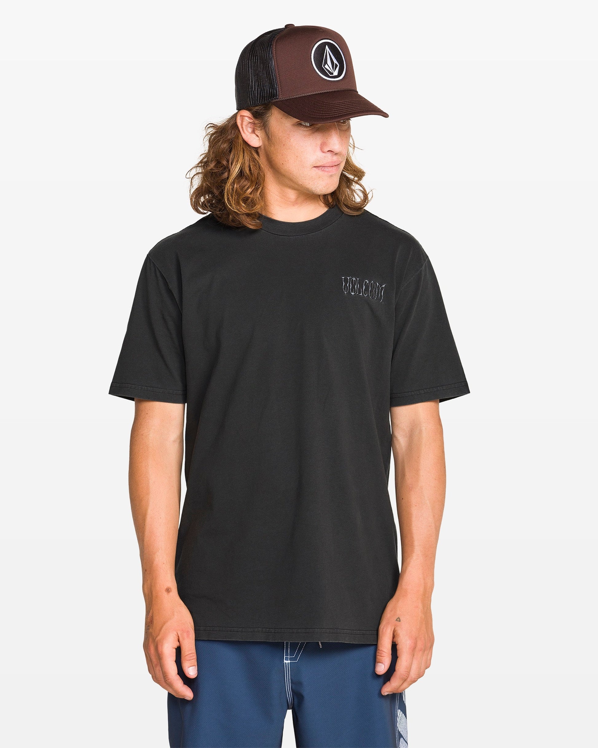 A man wearing the Volcom Men's Proto Type Short Sleeve Tee, blue shorts, and a black Volcom cap stands against a white background, looking down and to the side, channeling effortless style.