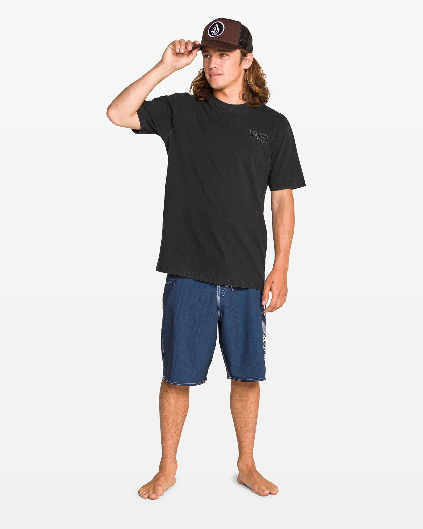 A young man wears the Volcom Men's Proto Type Short Sleeve Tee in black and blue board shorts, standing barefoot on a white background, touching his black cap—an effortlessly relaxed style ideal for pairing with Volcom embroidered sweatshirts or loose-fit crews.