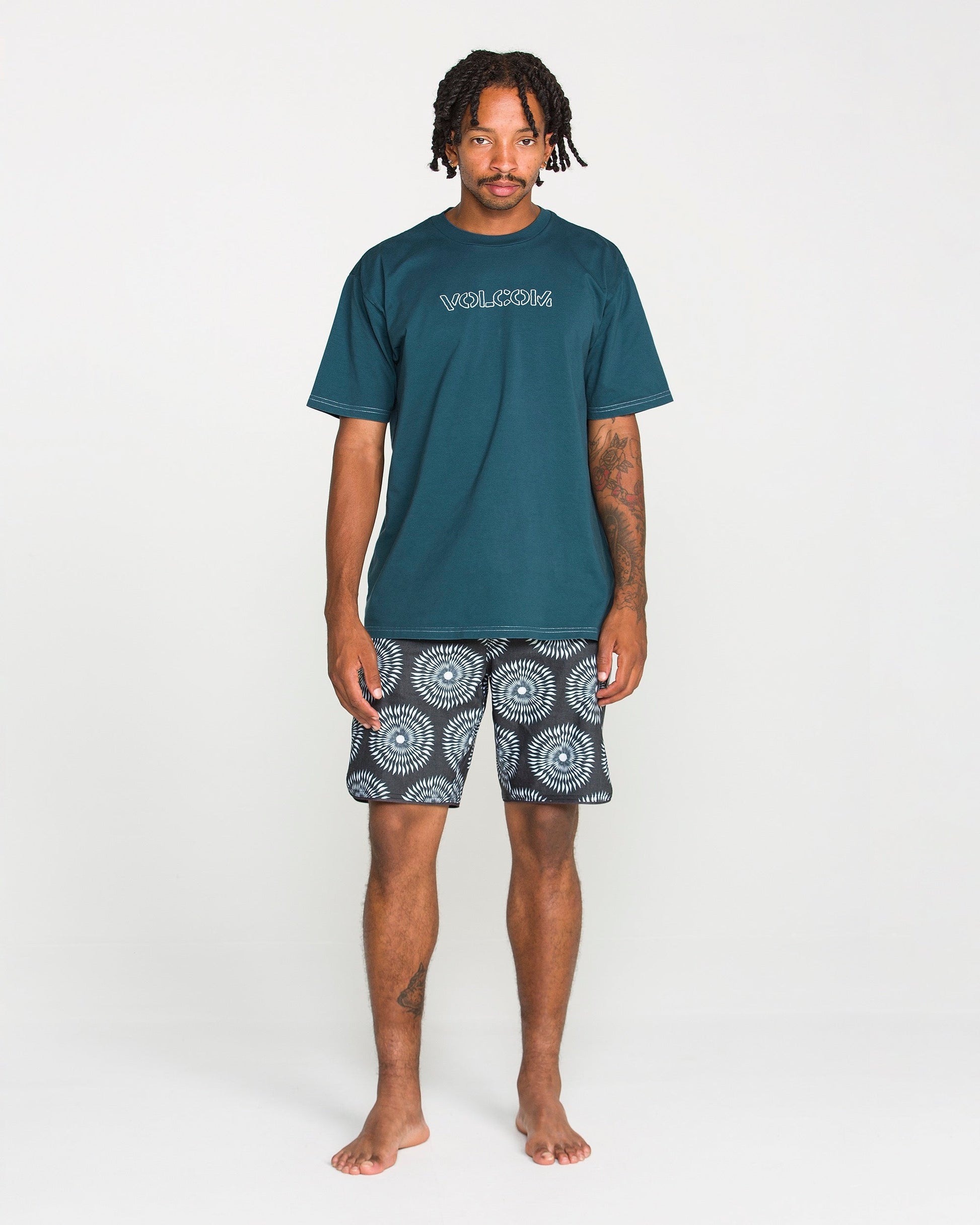 A man stands barefoot against a white background, wearing Volcom's Men's Crassy Short Sleeve Tee in teal—crafted from premium cotton with a soft-hand screenprint—and patterned black-and-white shorts.